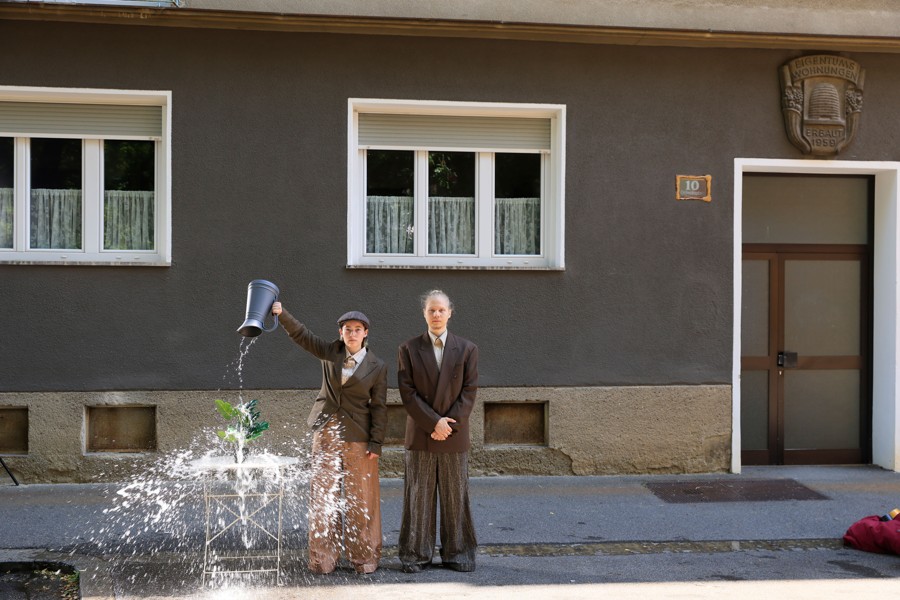 Ein Mann schüttet einem anderen Mann Wasser aus einer Kanne auf den Kopf; die beiden stehen vor einem Haus