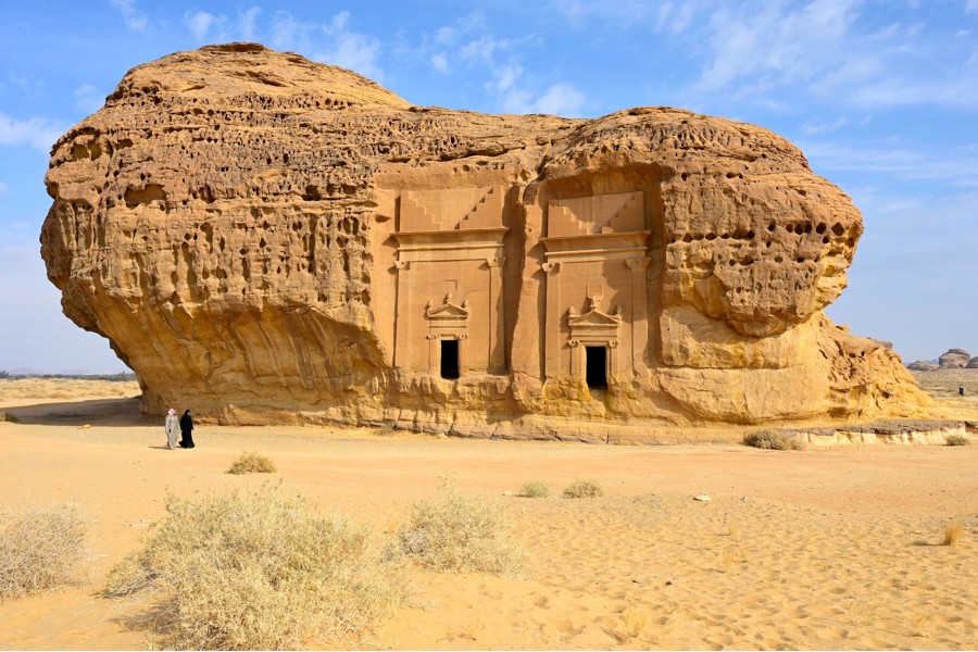 Zwei Araber vor riesiger Sandstein-Felsformation in der Wüste, in deren Seite 2 Eingangsportale zu erkennen sind